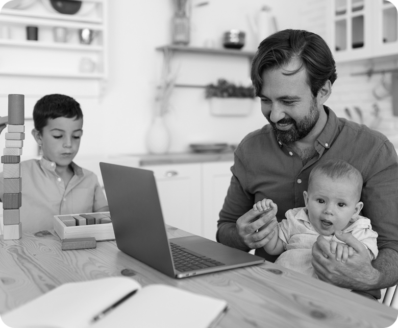 father with a beard sitting at a kitchen table, working on his laptop while holding his baby in his lap