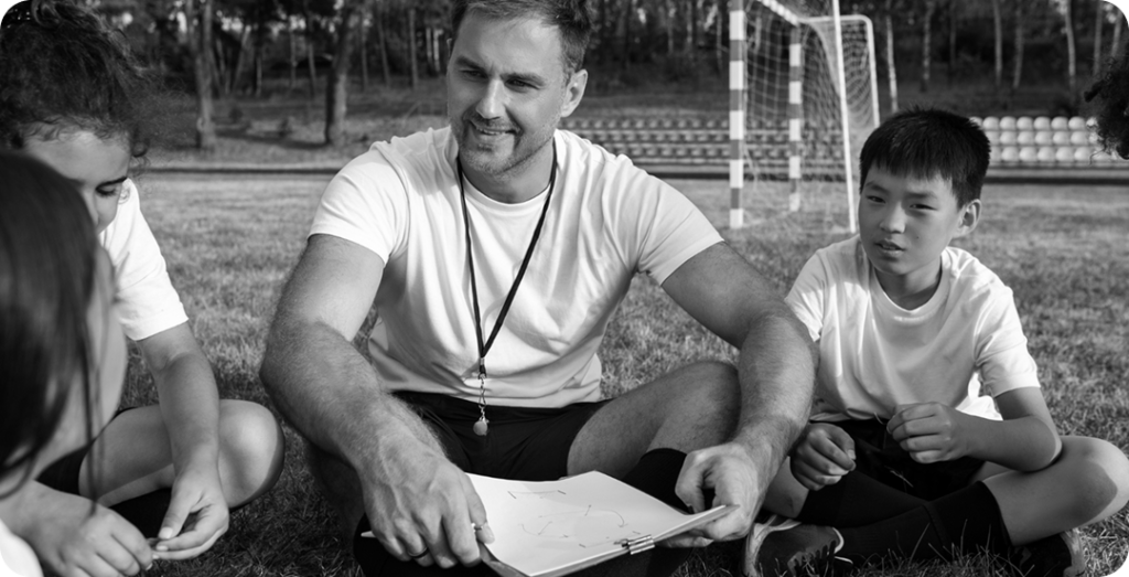 male soccer coach sitting on the grass with a group of young children. He is smiling and pointing to a diagram on a clipboard as he talks to them